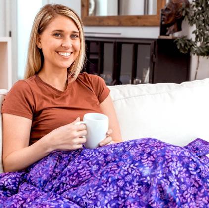 A blonde women resting under a weighted purple blanket holding a white mug.